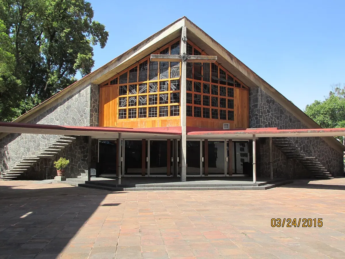 A modern church with a steep A-frame roof, a large window and a tall cross in front, on a brick plaza