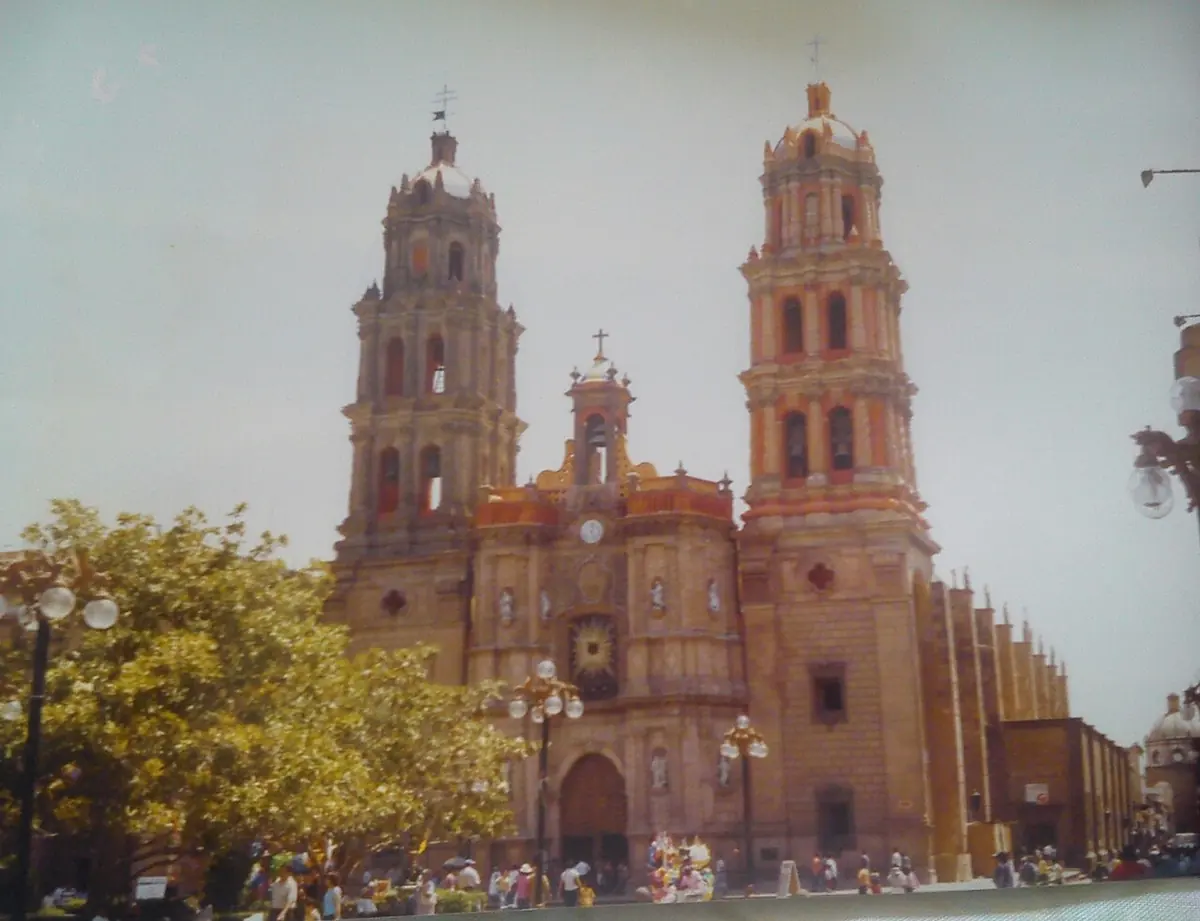 A pink stone baroque cathedral with two tall bell towers, seen across a plaza with trees