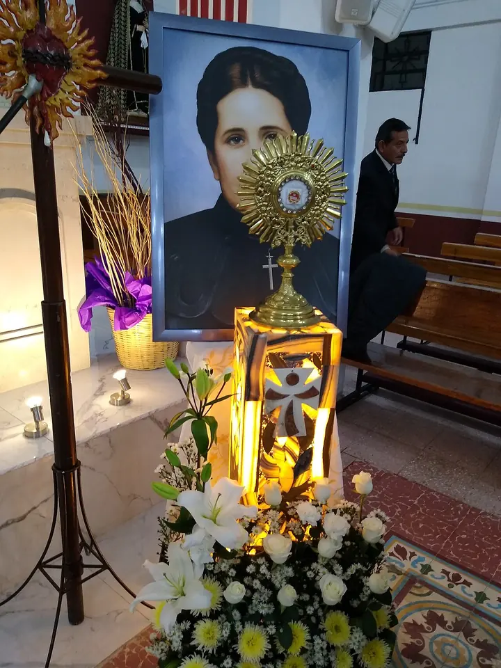 A gold reliquary on a stand surrounded by white lilies, in front of a framed portrait of a dark-haired woman