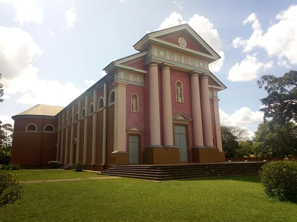 The pink and red brick facade of St. Joseph's Cathedral in Gulu, with tall columns and a triangular pediment, set on a green lawn under a blue sky