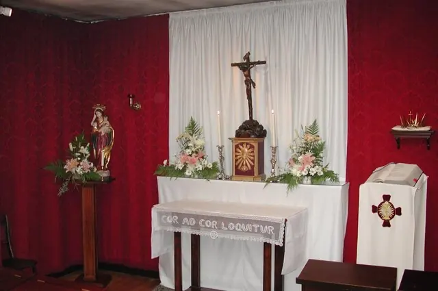 A small chapel with red walls, a white altar with a crucifix and flowers, and an altar cloth embroidered with the words Cor ad cor loquitur