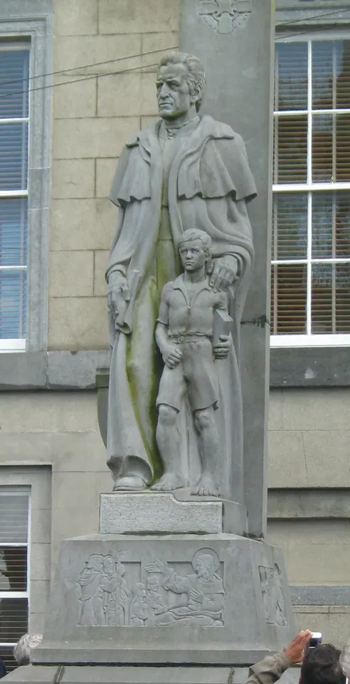 Stone statue of Edmund Rice in a long coat with his hand on the shoulder of a schoolboy, on a carved pedestal in a town street