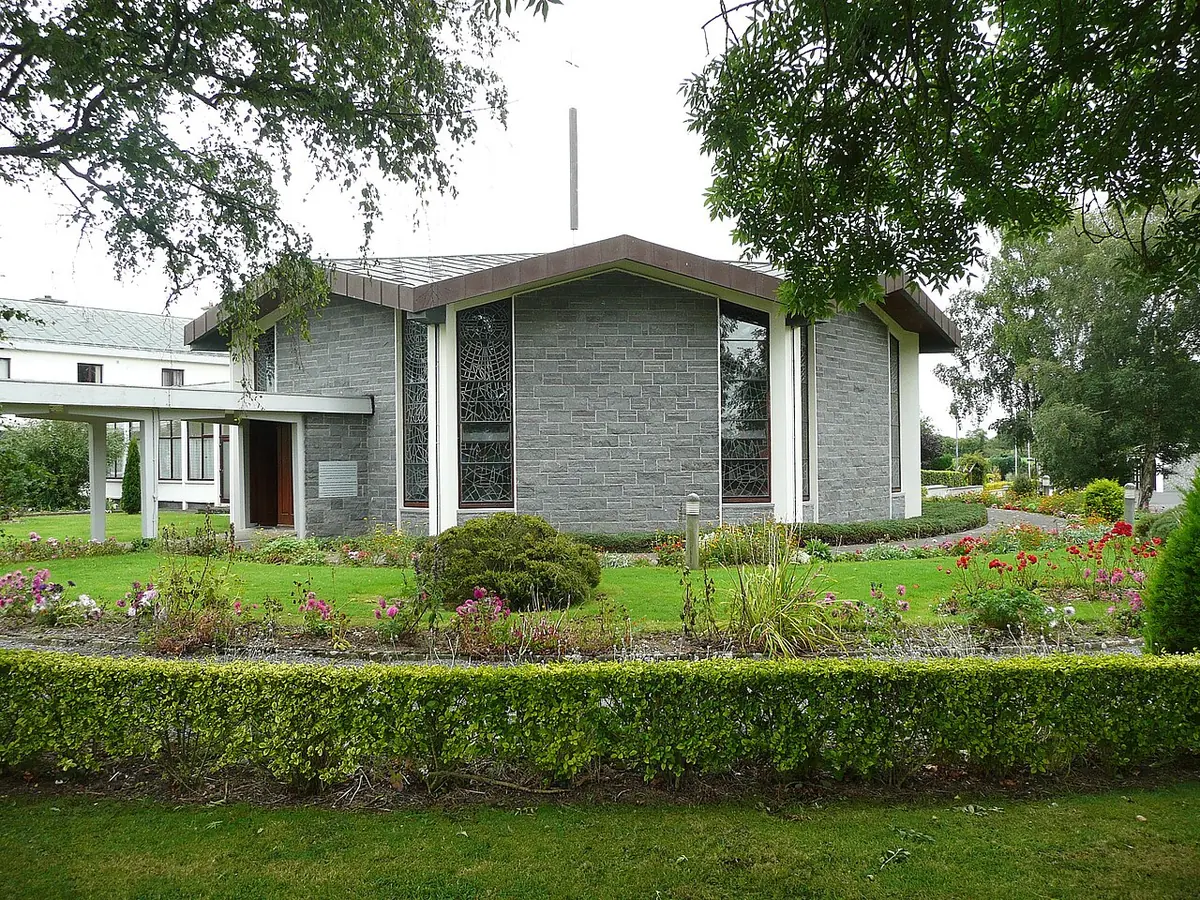 Modern gray stone chapel with tall stained-glass windows behind flower beds and a clipped hedge