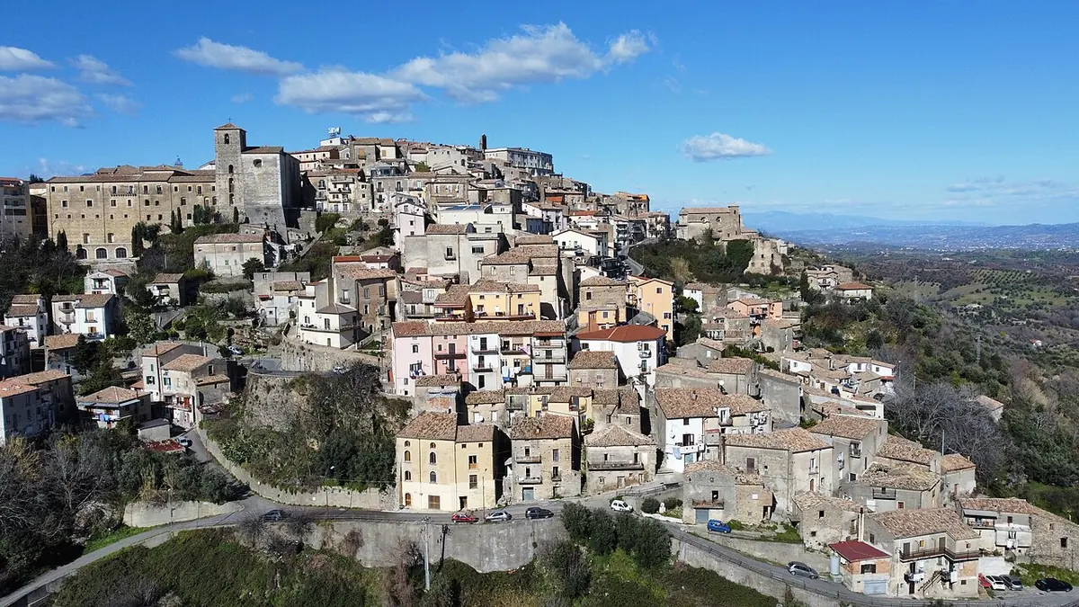 Aerial view of a hill town of pale stone houses crowded around a church and bell tower, with green hills and the sea in the distance