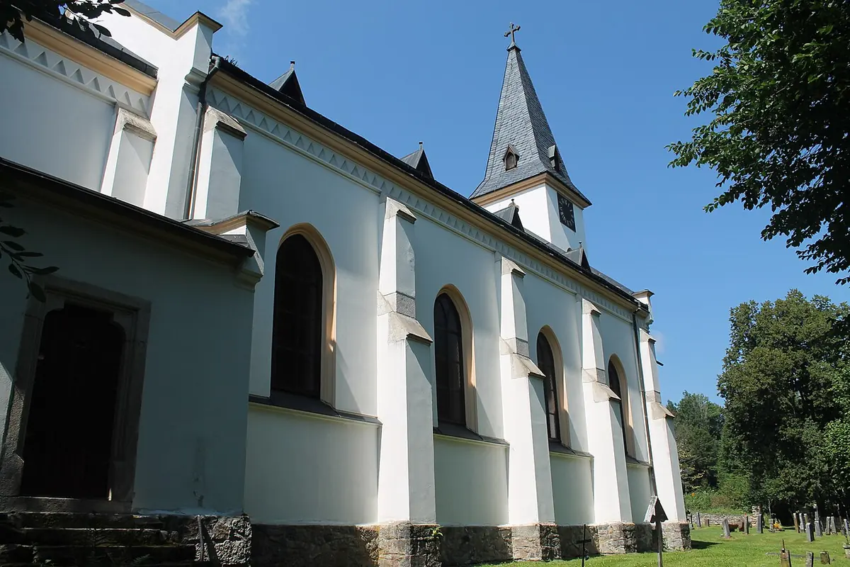 White village church with tall arched windows and a slate-roofed bell tower among trees, with a small graveyard beside it