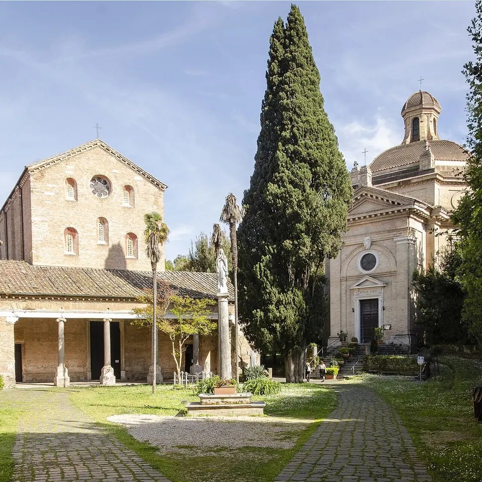 Brick Romanesque abbey church with a rose window and a portico, beside a tall cypress and a domed church, around a grassy courtyard
