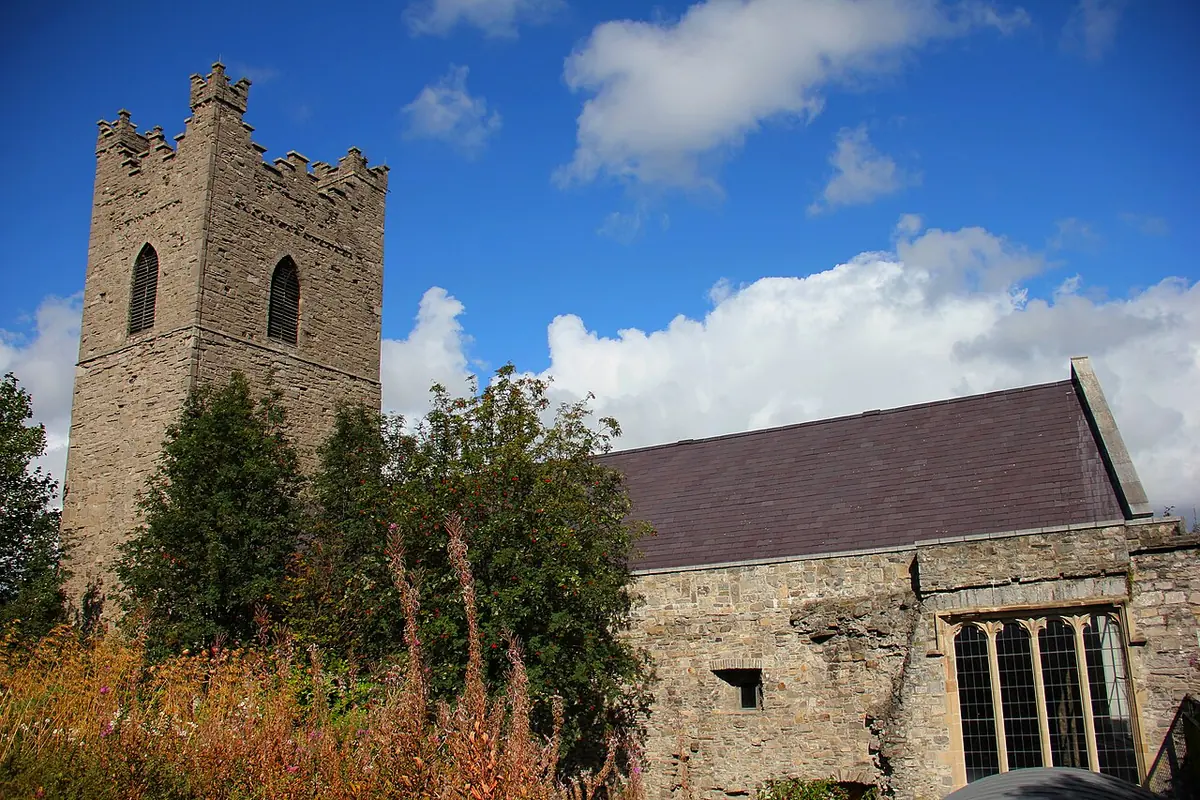 A medieval stone church with a square battlemented tower and a long slate roof, seen over trees and wild plants under a blue sky