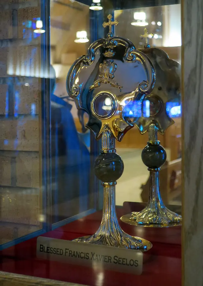 An ornate gold reliquary with a small round relic window, standing above a framed holy card with a photograph of the priest