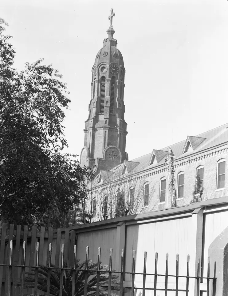 Black-and-white photograph of the tall ornate brick bell tower of St. Mary's Assumption Church rising above trees and a long brick building behind a fence
