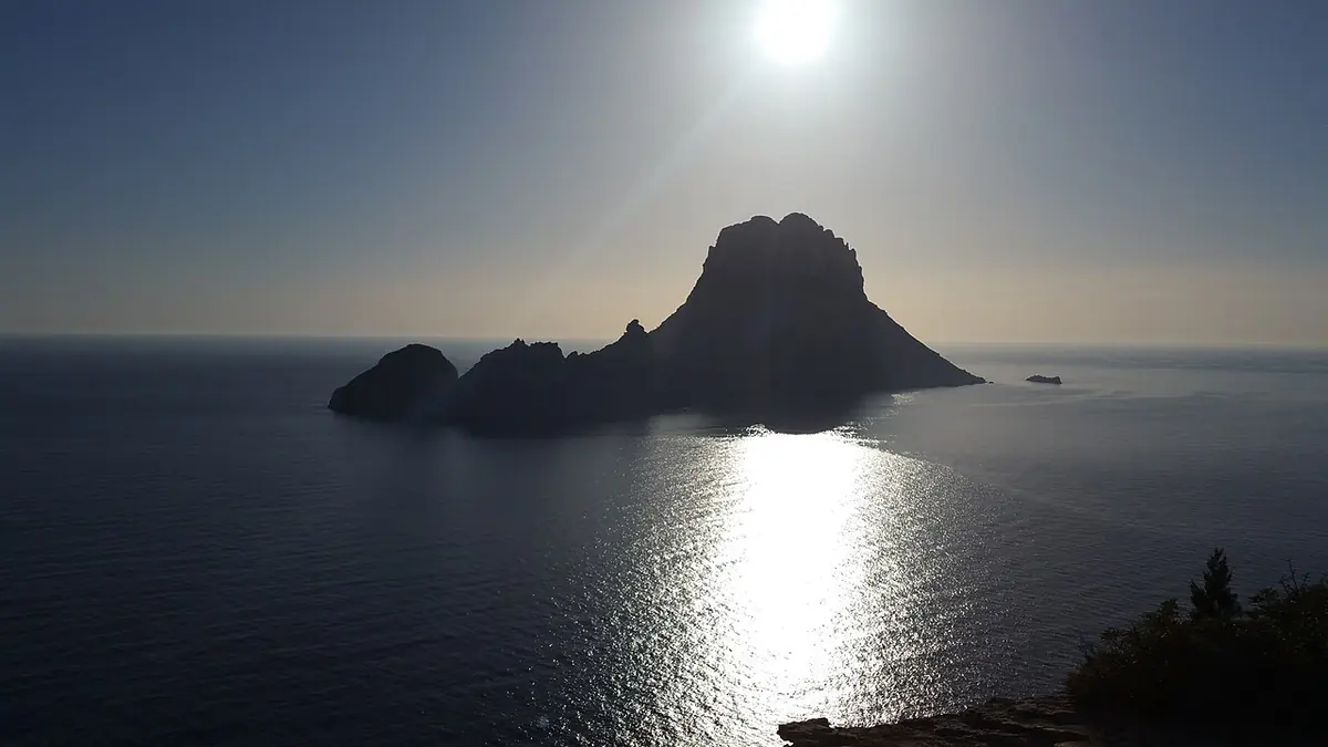 The steep rocky islet of Es Vedrà and its smaller neighbor silhouetted against a bright sun over a shining sea