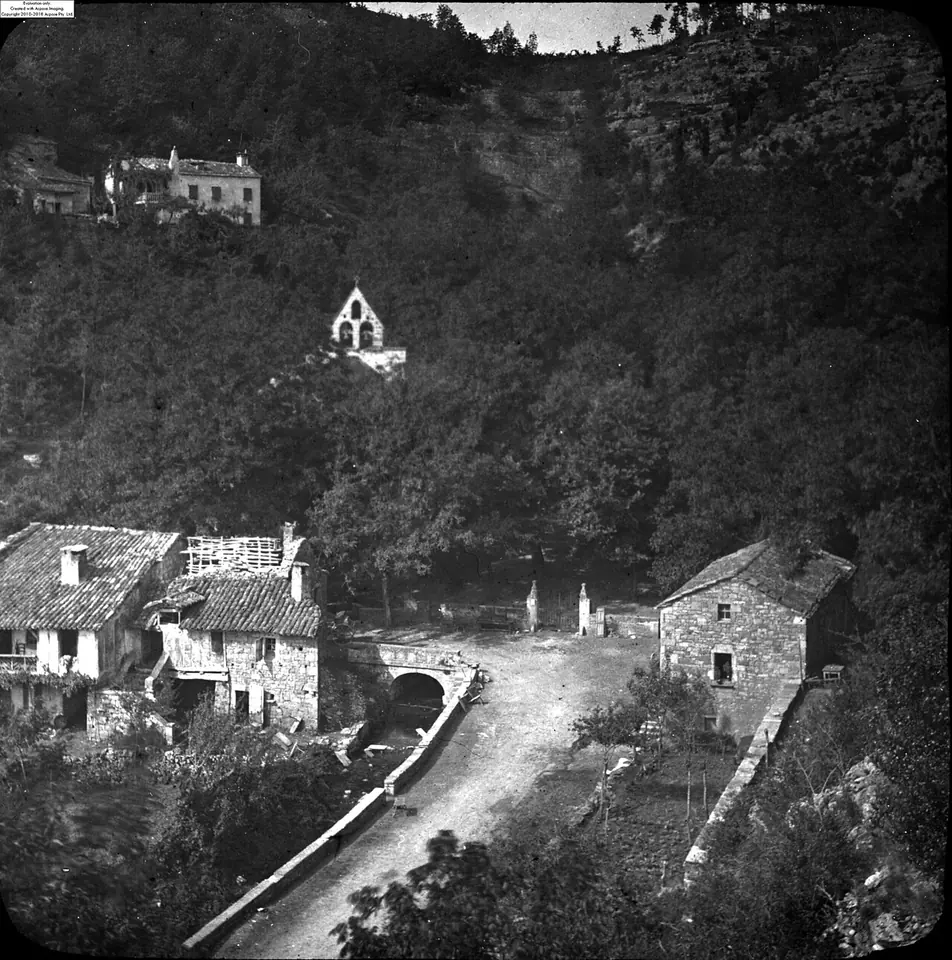 Old black-and-white photograph of a wooded gorge with a small chapel on the slope, stone houses and a road along a stream below