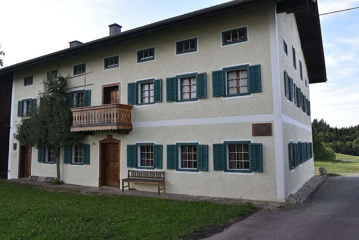 Long white two-story Austrian farmhouse with green shutters, a wooden balcony and a bench outside, on a green lawn