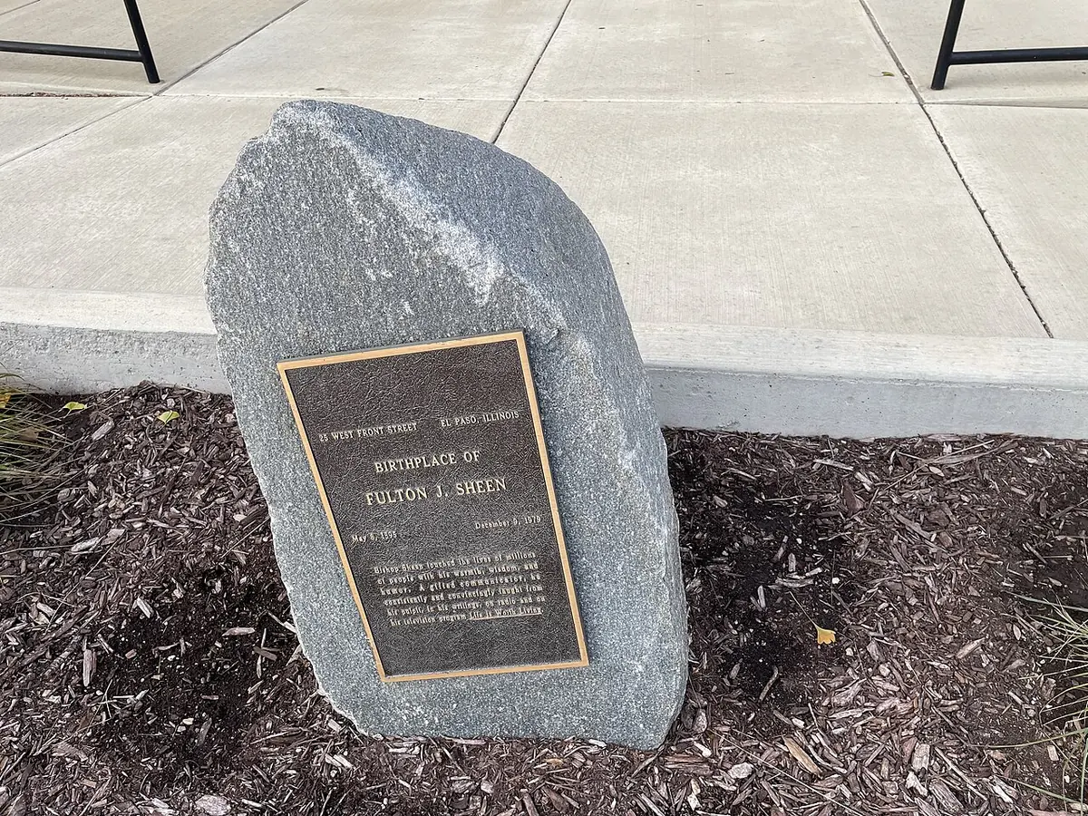 A rough gray boulder set in mulch beside a sidewalk, with a bronze plaque reading Birthplace of Fulton J. Sheen