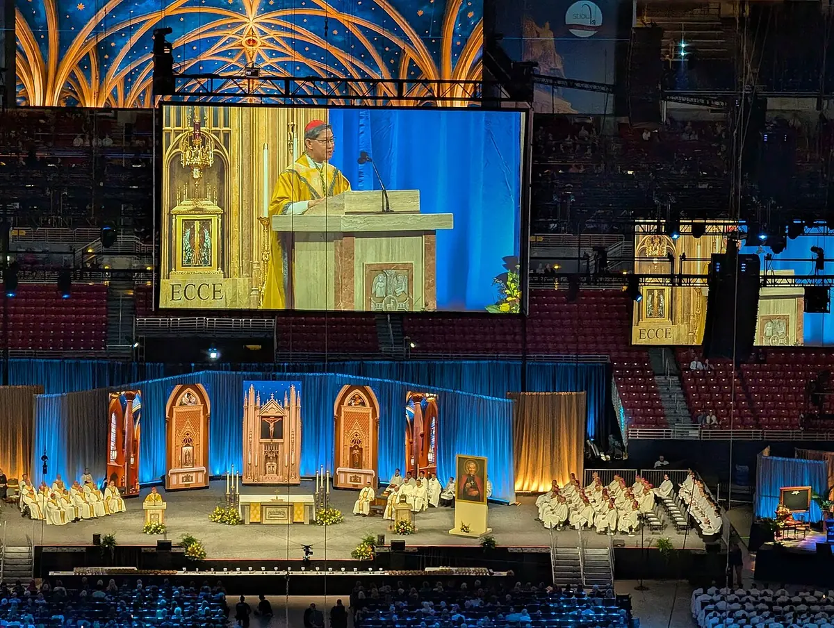 A cardinal in gold vestments at an ambo shown on a giant screen above the altar stage, with bishops and priests seated around it