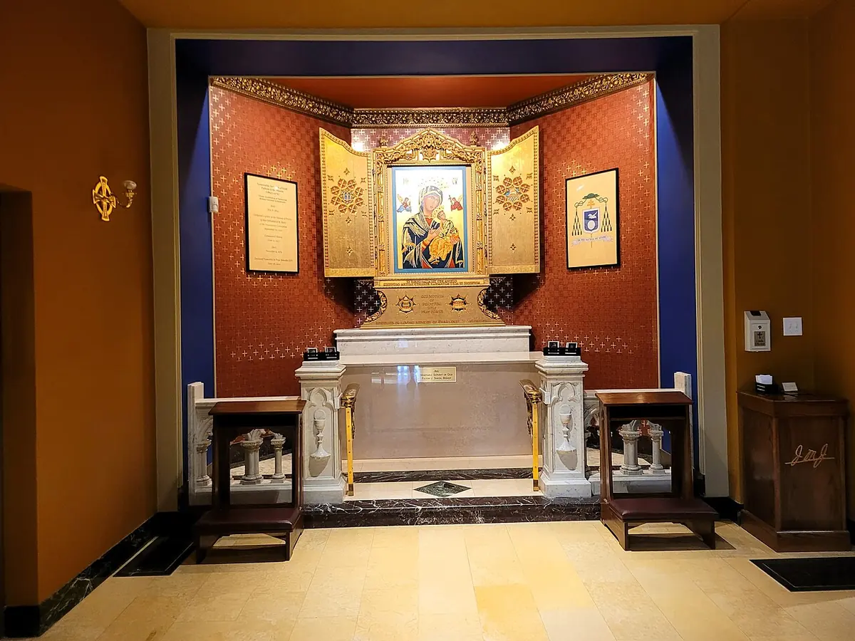 A side chapel with red walls where a white marble tomb stands beneath a gold-framed icon of Our Lady of Perpetual Help, with kneelers on either side