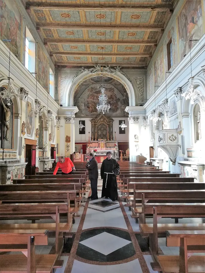 The long nave of a church with a painted wooden ceiling, white walls with frescoes, wooden pews and a marble altar at the far end, with two friars walking in the aisle