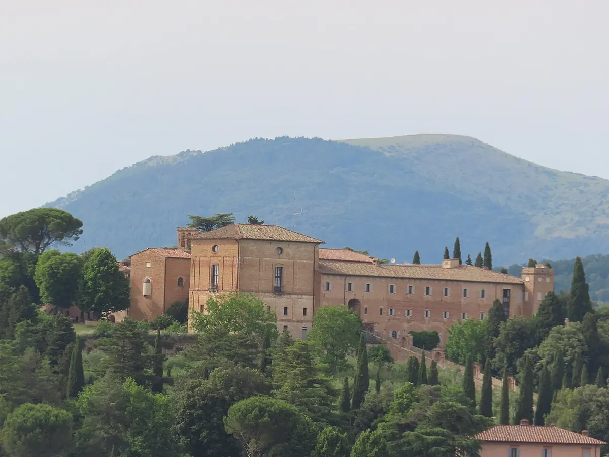 A long brick friary building on a wooded hilltop among cypress and pine trees, with mountains behind