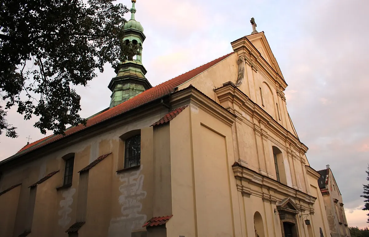 Cream-colored Baroque church with a red tile roof, buttressed side walls and a green onion-domed bell tower, seen from a corner at dusk
