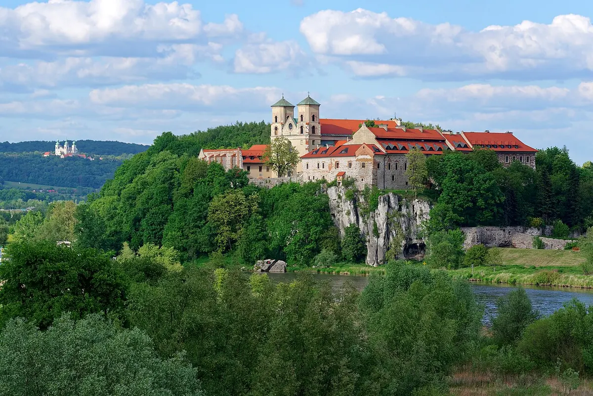 A white abbey with twin towers and red roofs on a limestone cliff above a river, surrounded by green trees under a blue sky with white clouds