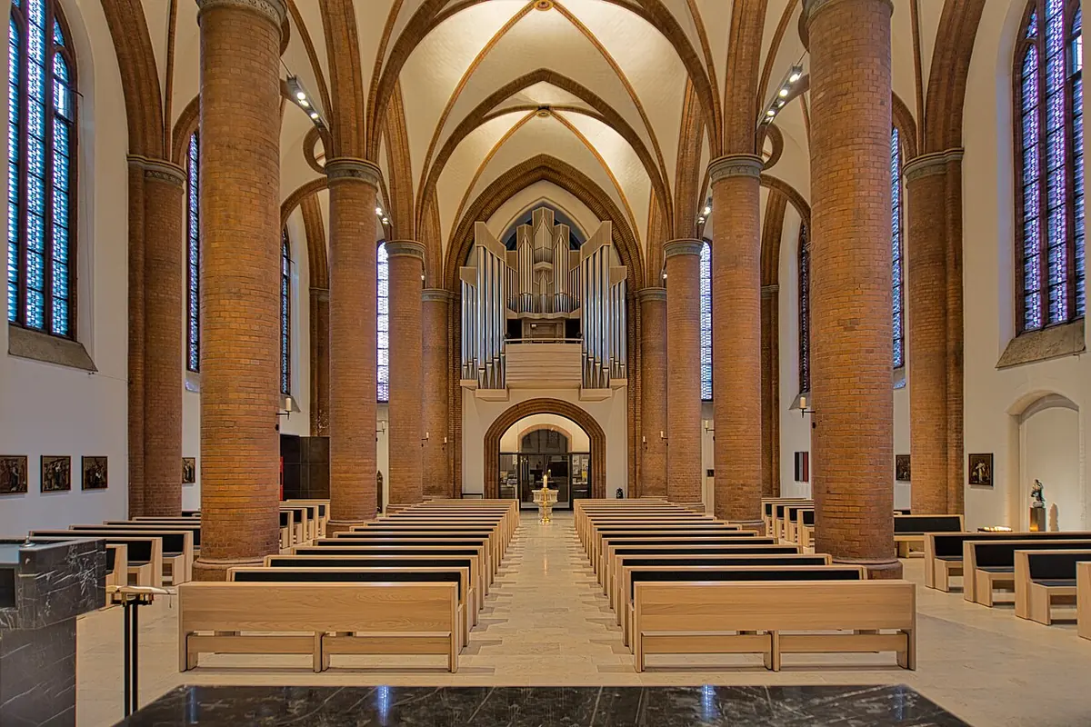 The interior of a brick Gothic Revival church with tall round pillars, pale wooden pews and the organ loft at the far end