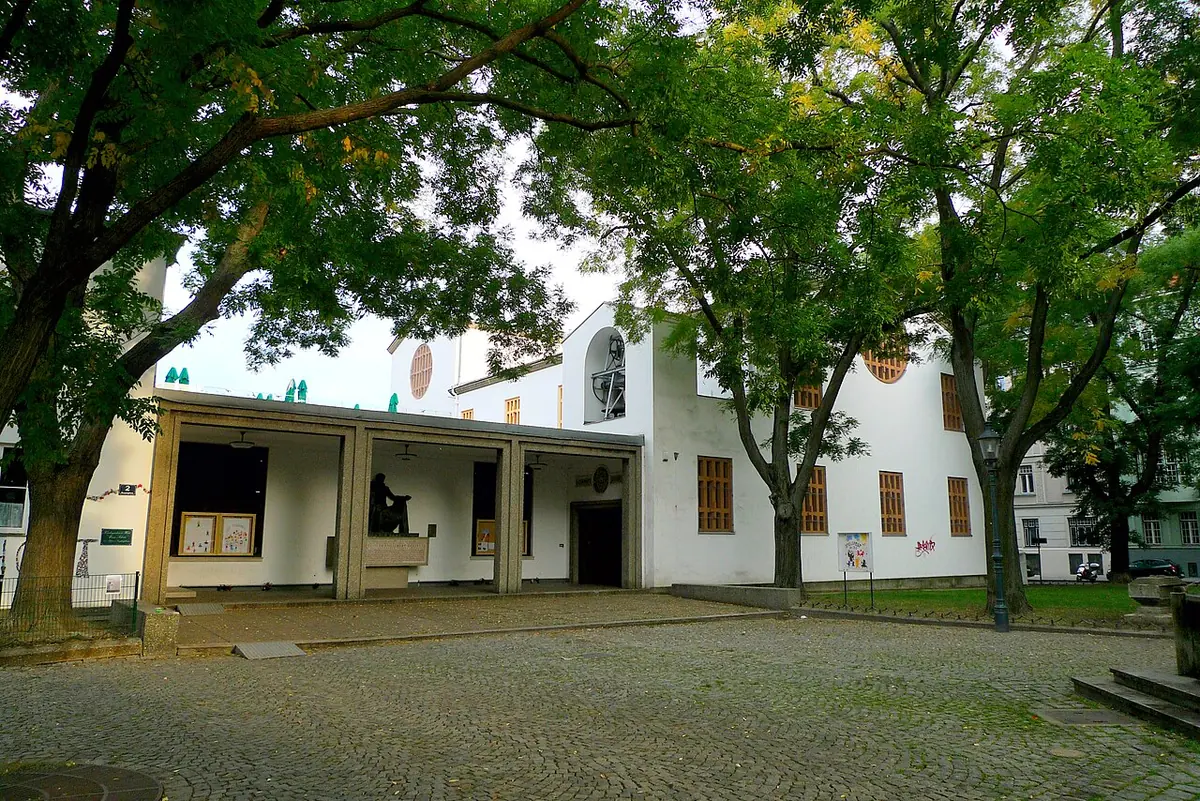 Low white church with a covered porch and a round window, behind a cobbled square shaded by large trees