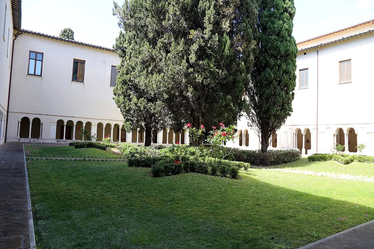 A quiet cloister garden with lawns, clipped hedges and tall cypress trees, surrounded by a low arcade of small columns