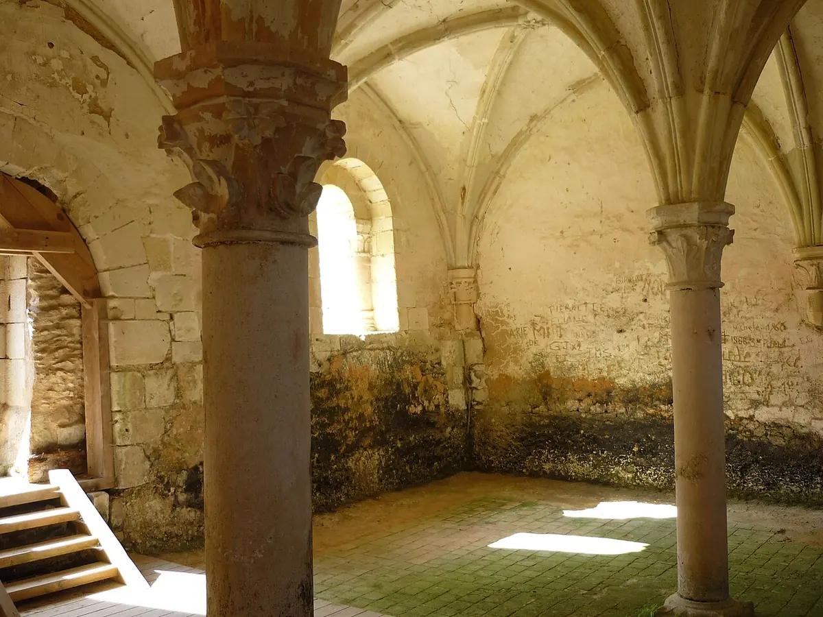 A small stone hall with ribbed Gothic vaults resting on two slender columns, sunlight falling across a worn tile floor