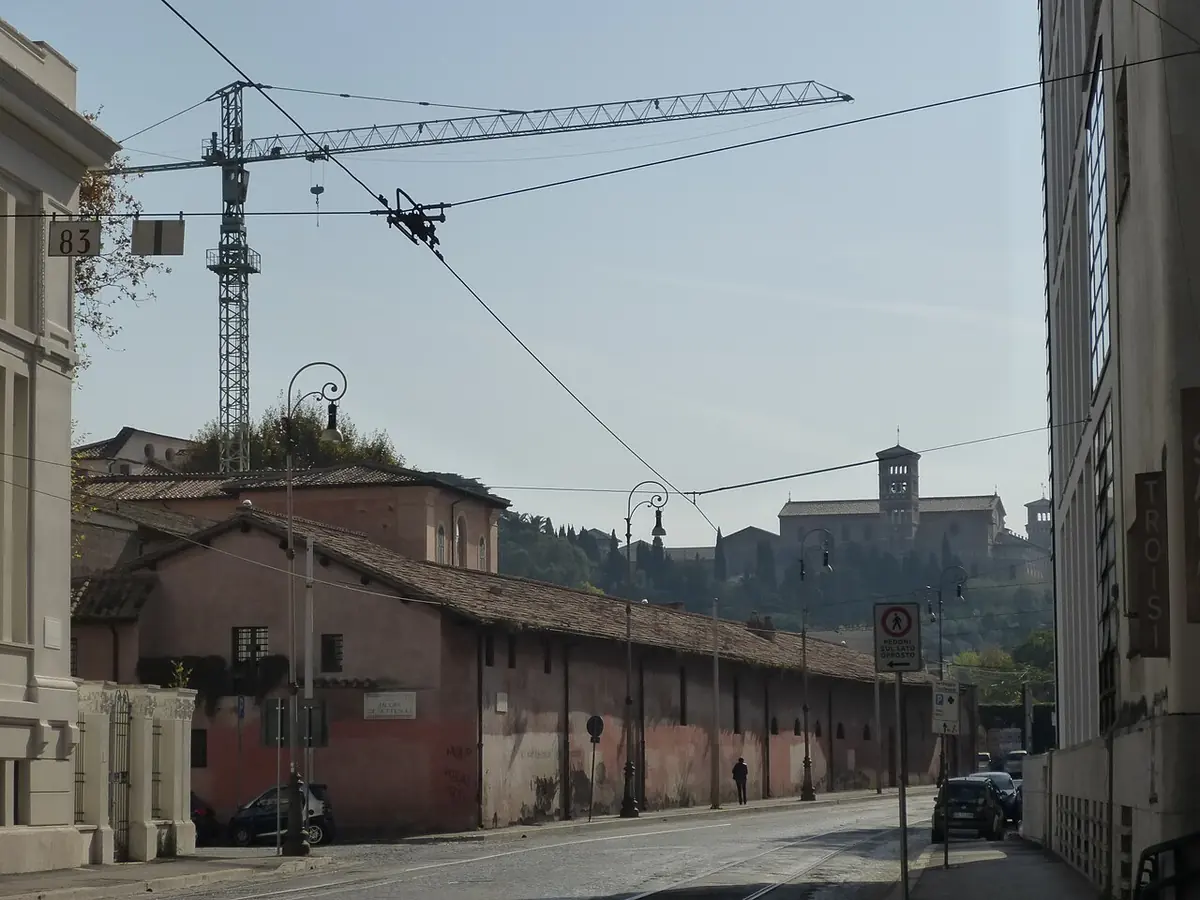 A Roman street running along a long low wall with old buildings, with a bell tower beyond and tram wires overhead