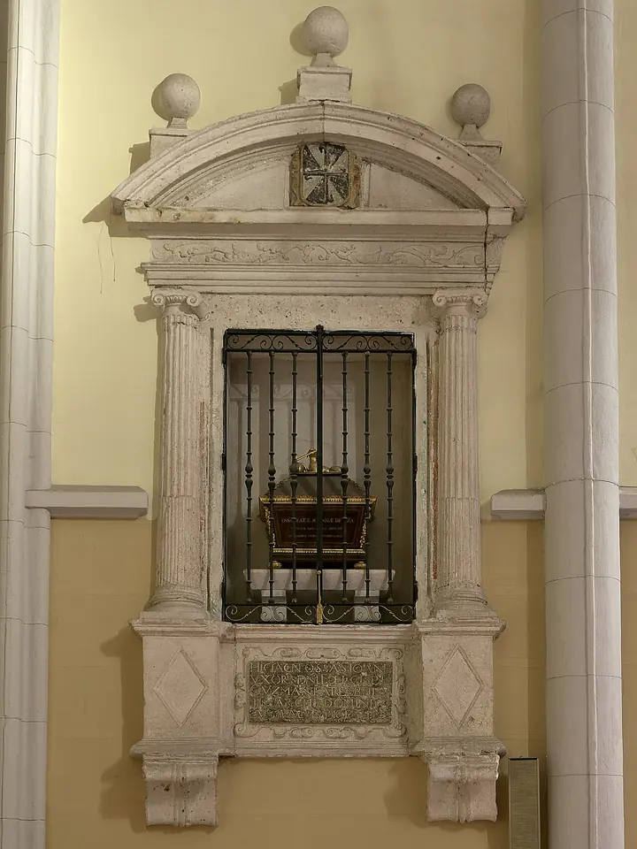 A gilded reliquary chest behind an iron grille in a white stone wall niche framed by columns and a pediment