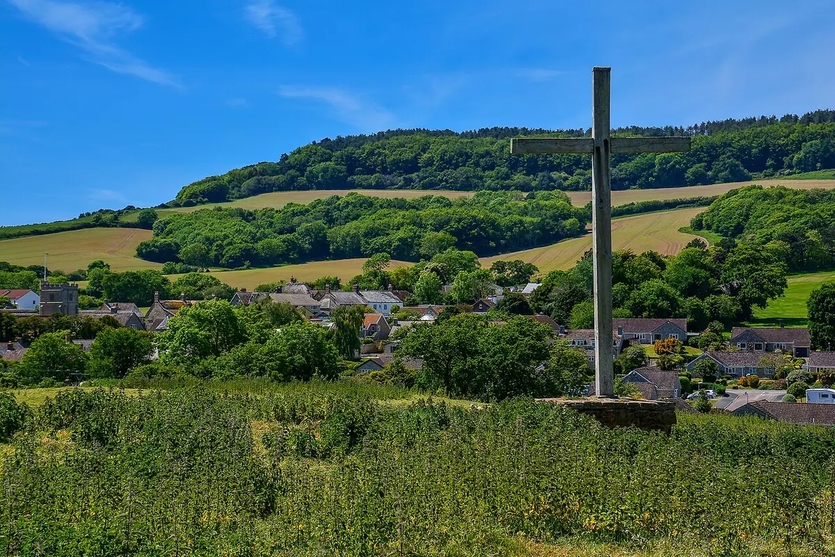 A tall dark wooden cross standing in a grassy field above the village of Chideock, with wooded green hills behind