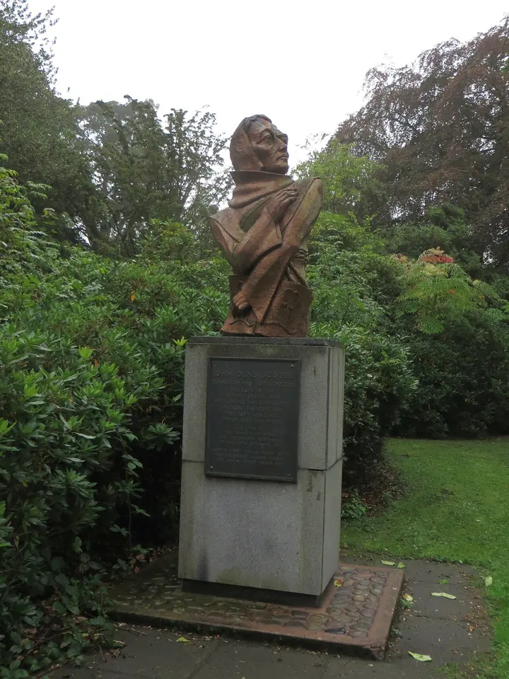 A bronze bust of a hooded friar with his arms folded, on a gray stone plinth with a plaque, among trees and shrubs in a park