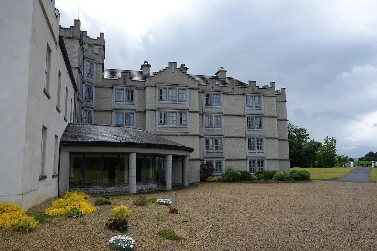 A long gray castellated college building with many windows behind a gravel drive and flower beds under a cloudy sky