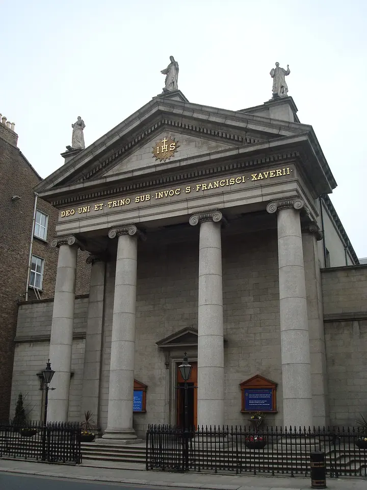 The classical stone portico of a Dublin church with four columns, a pediment bearing the IHS emblem and statues on the roofline