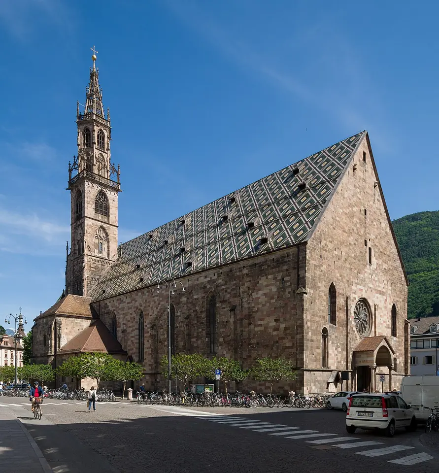 A Gothic sandstone cathedral with a tall openwork bell tower and a steep roof of colored diamond-patterned tiles, seen from a street corner with bicycles in front