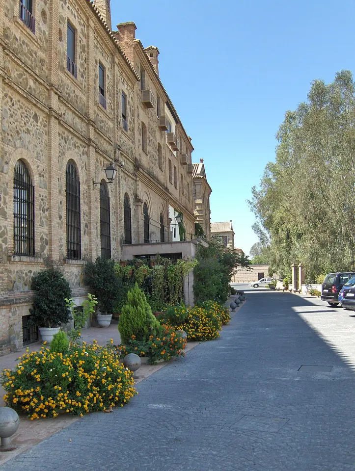 The long stone-and-brick facade of a large seminary building with arched windows, flowering shrubs along a paved lane