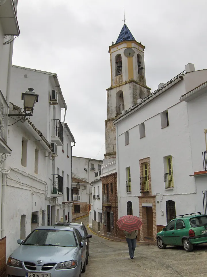 A narrow street of white houses climbing toward a church bell tower with a blue-tiled spire, a person with a red umbrella walking up it