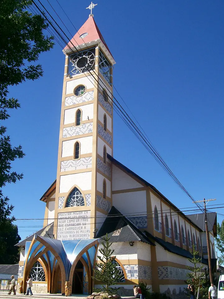 Tall church with a white and ocher clock tower and a pointed red roof against a deep blue sky, with a modern arched entrance porch