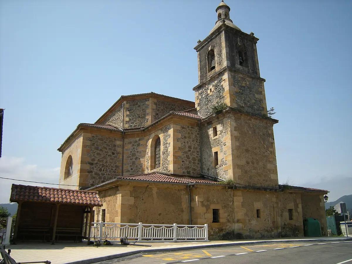 A sandstone country church with a square bell tower and a red tile roof beside a quiet road, with a covered porch along its side