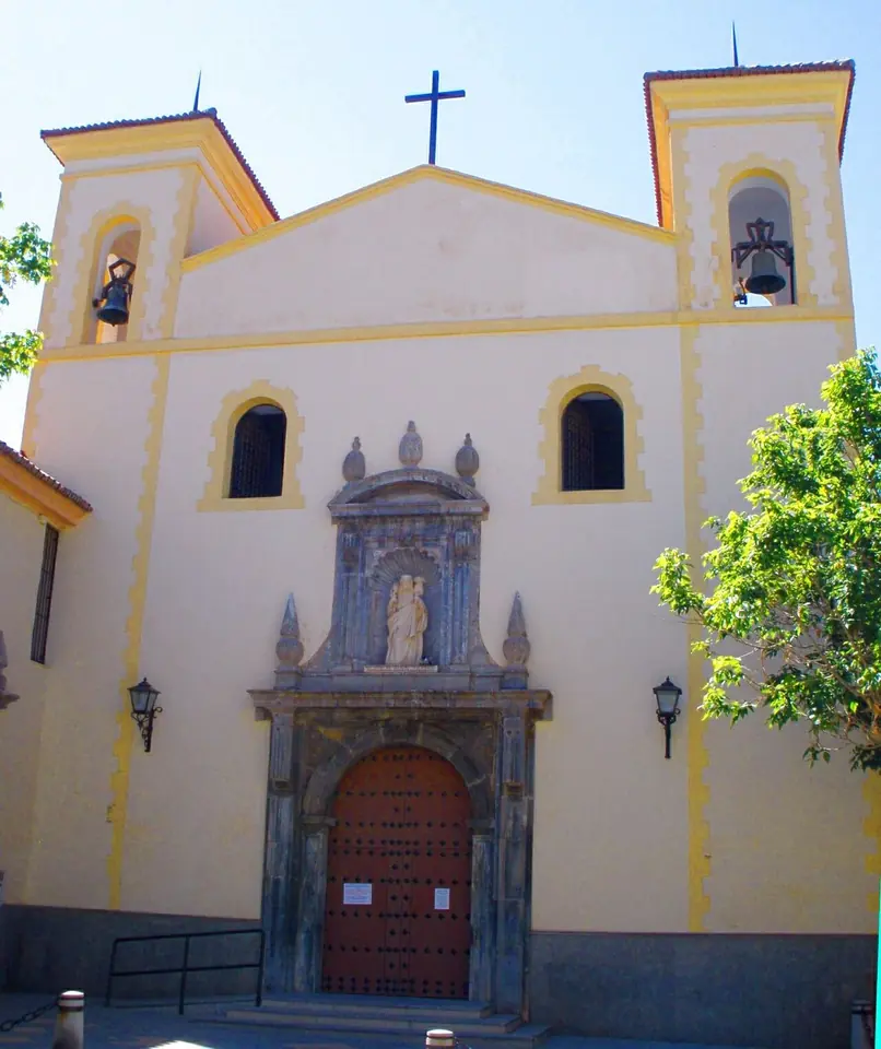 The cream-and-yellow facade of a church with two small bell towers, a cross on top and a carved stone doorway with a statue in a niche