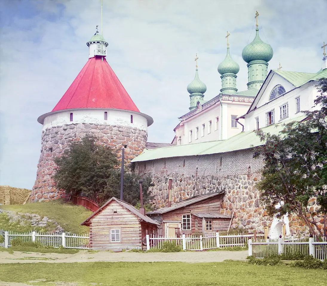 An early color photograph of a round stone tower with a red conical roof beside a white monastery church with green onion domes