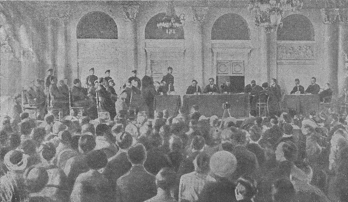 A black-and-white photograph of a crowded hall with chandeliers and arched windows, the judges at a long table at the far end and spectators filling the floor