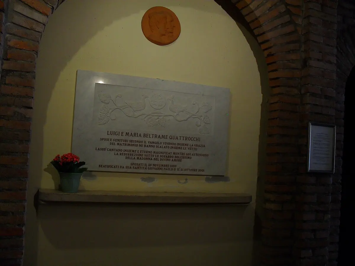 A marble tomb slab with an inscription set into a brick arch in the crypt of the Shrine of Divine Love, with a red flowerpot on a ledge below