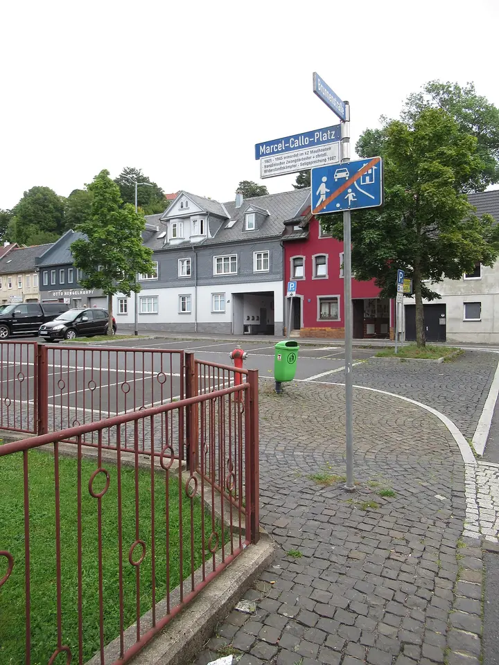 A small town square with cobblestones, a red railing and a blue street sign reading Marcel-Callo-Platz, with houses behind