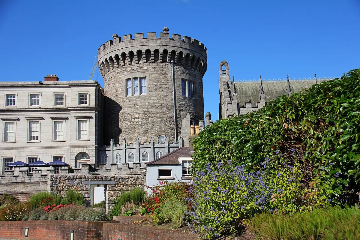A round medieval stone tower with battlements, joined to later Georgian buildings, seen over a garden of shrubs and flowers