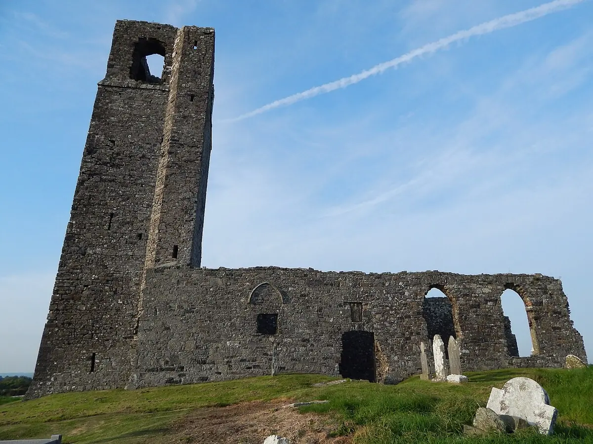 A ruined medieval stone church with a tall square tower on a green hilltop under a blue sky, with old gravestones in the grass