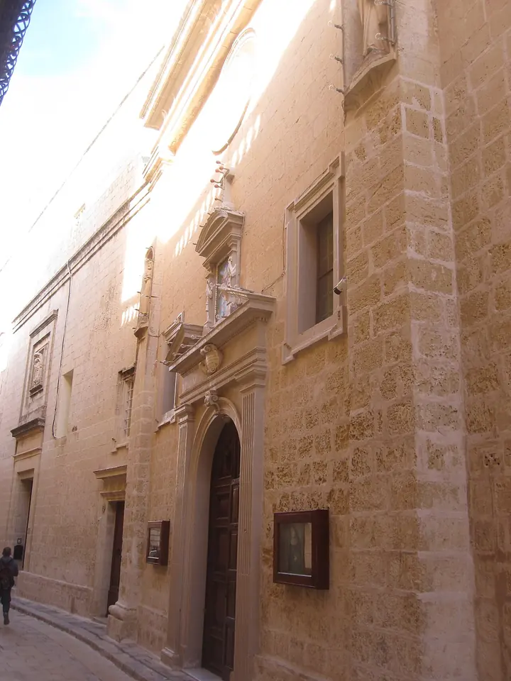 A narrow street of pale limestone in Mdina with the tall, plain facade of a monastery church and its doorway