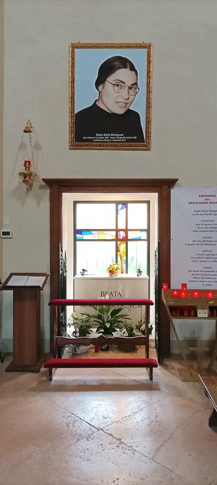 A small side chapel with a stone tomb under a window, red kneelers before it, and a framed photograph of a woman with glasses on the wall above