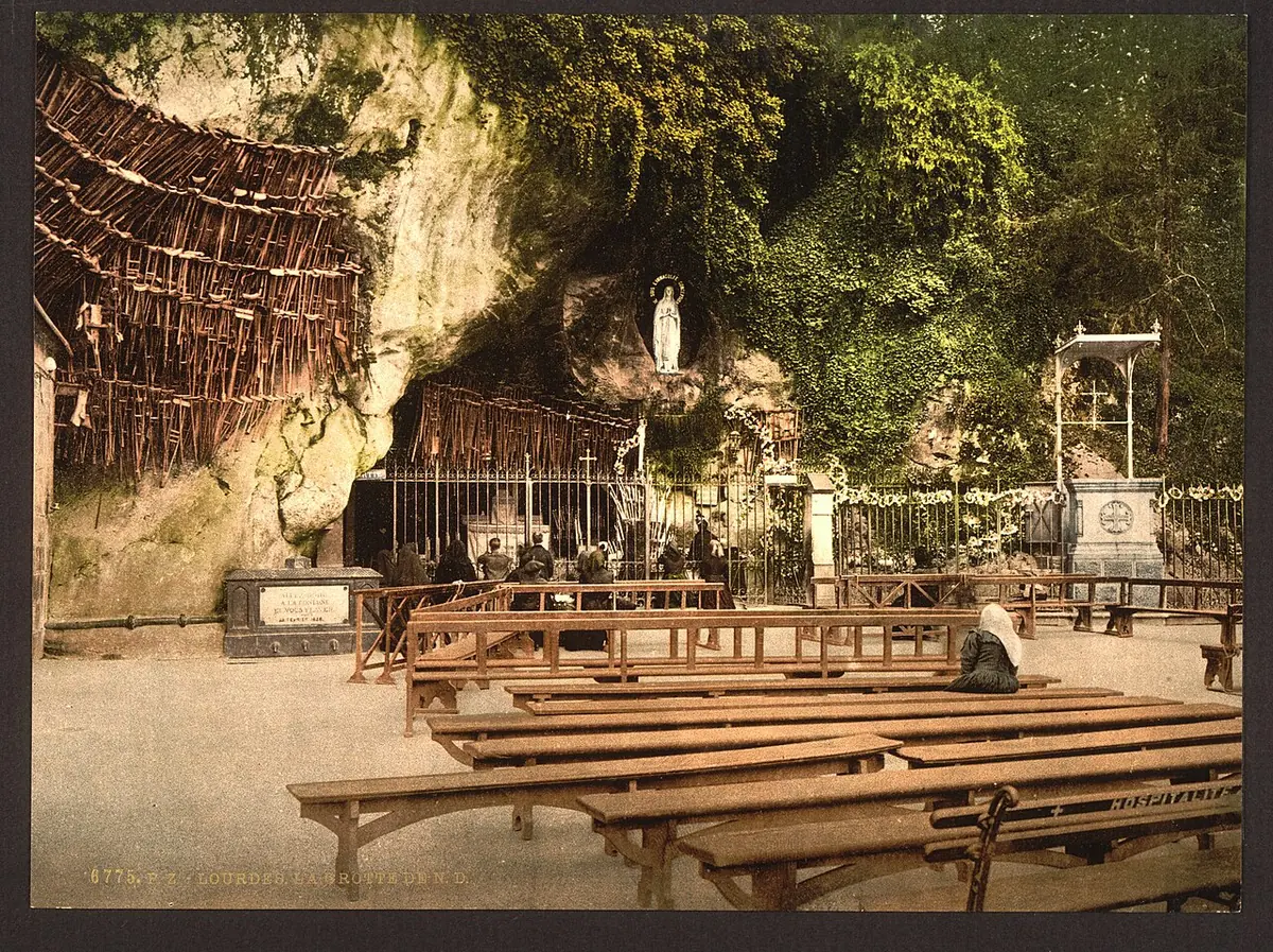 A tinted old photograph of the grotto at Lourdes with the statue of Our Lady in a niche of the rock and rows of wooden benches before it