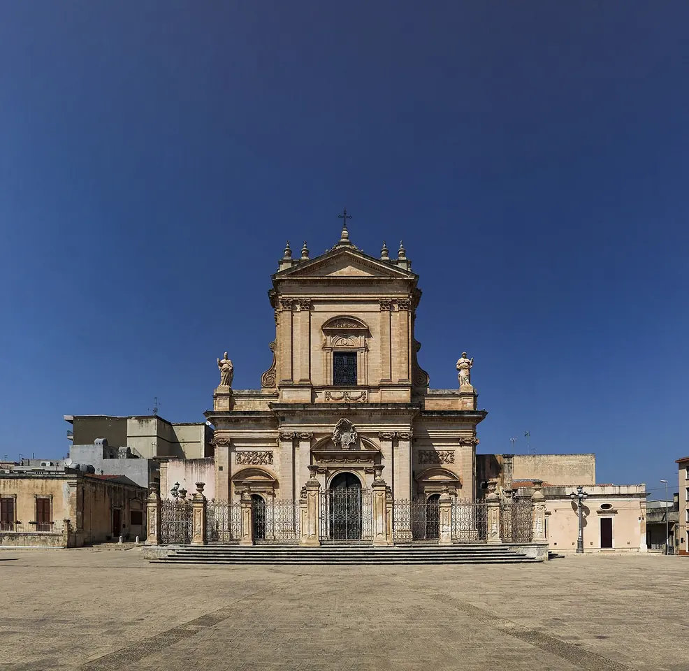 The golden stone Baroque façade of the Basilica of Santa Maria Maggiore in Ispica, Sicily, above a wide paved square under a deep blue sky