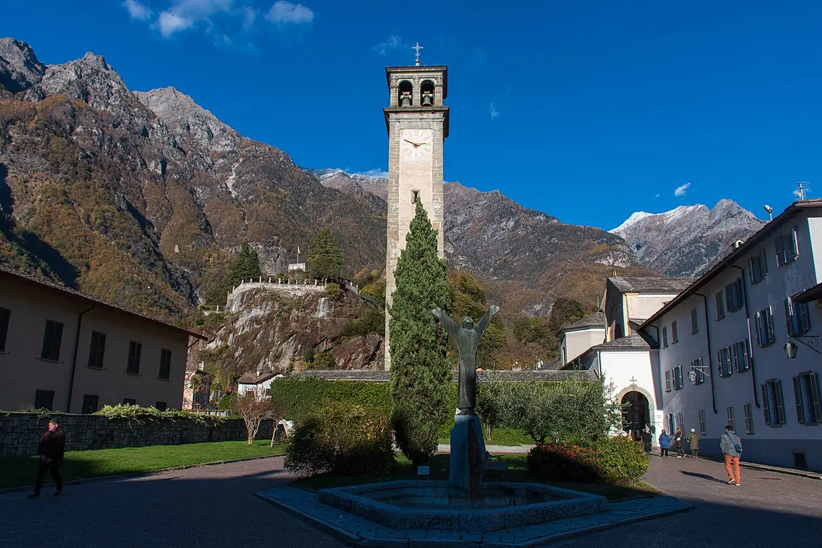 The tall bell tower of the Collegiate Church of San Lorenzo in Chiavenna, beside a garden and old buildings, with rocky mountains behind under a blue sky